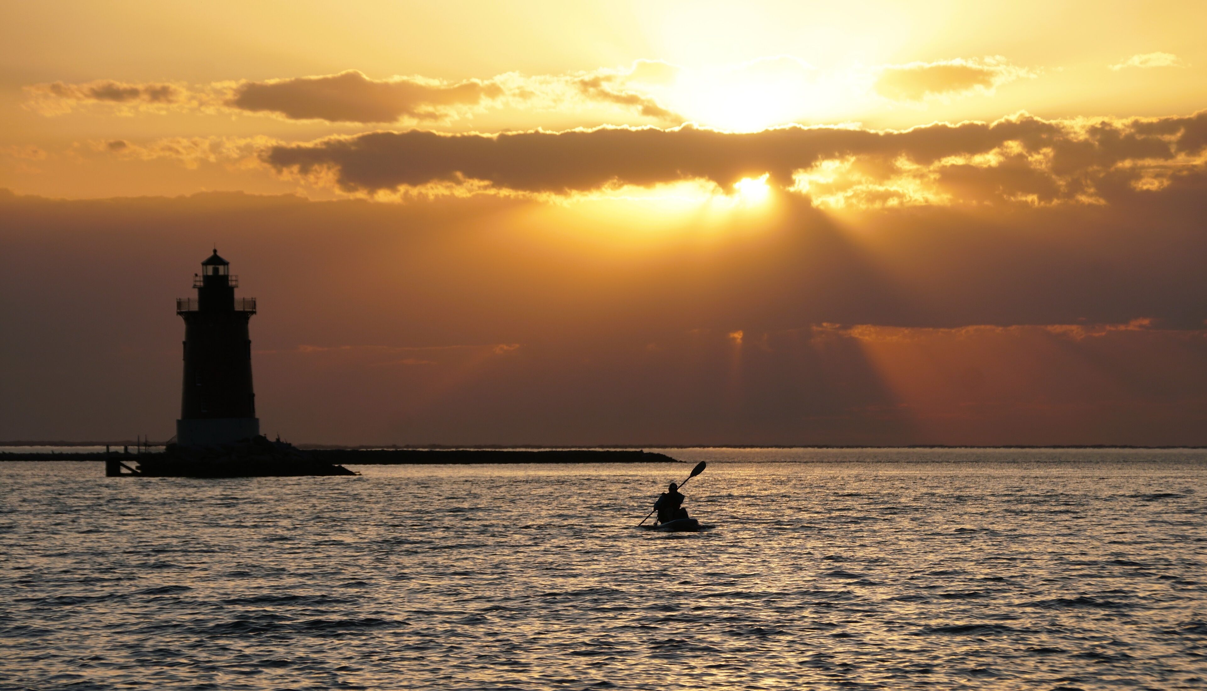 A lighthouse silhouette at sunset with rays of sunlight piercing through clouds, and a person paddling a kayak in the foreground.