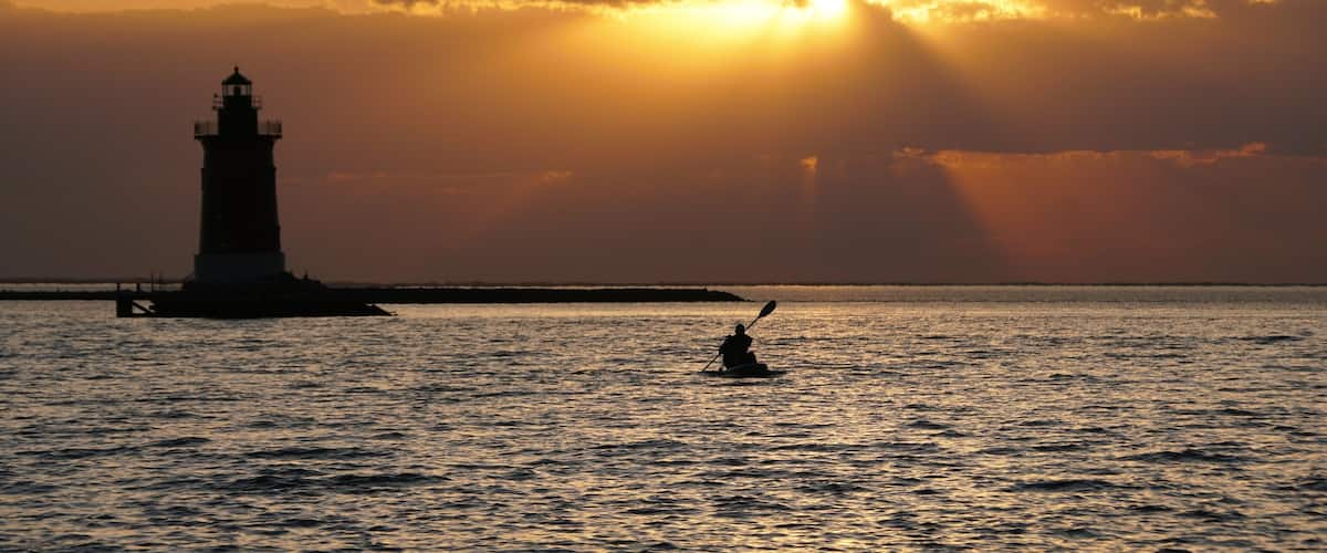 A lighthouse silhouette at sunset with rays of sunlight piercing through clouds, and a person paddling a kayak in the foreground.