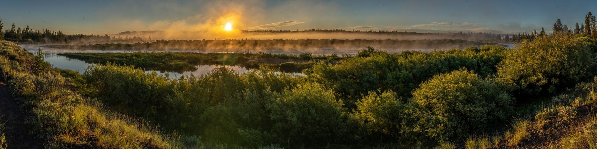 A sunrise over a forested landscape with a misty river.