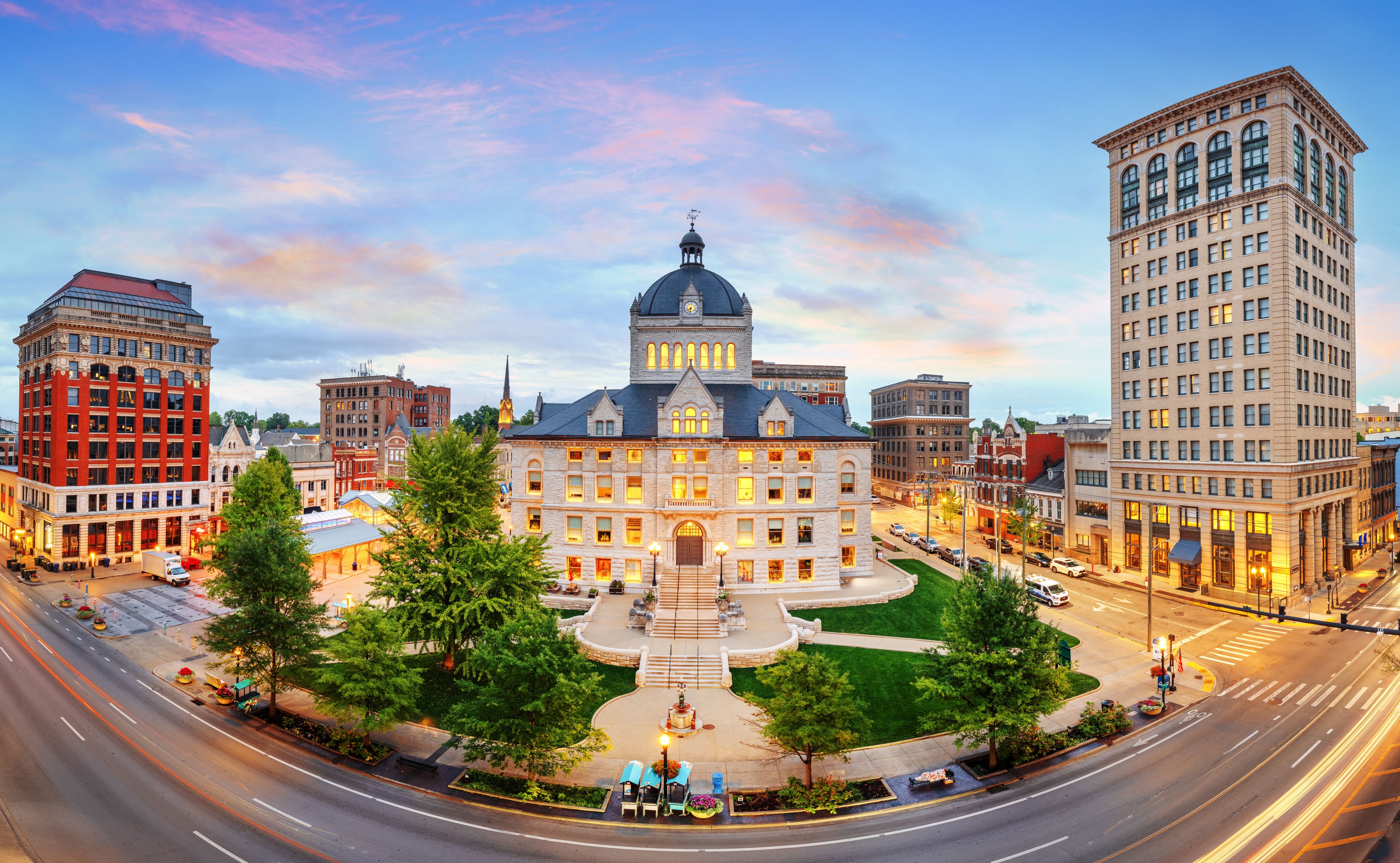 Un edificio histórico con una cúpula central y una torre del reloj, rodeado de modernos rascacielos y una plaza bien cuidada con árboles y una fuente.