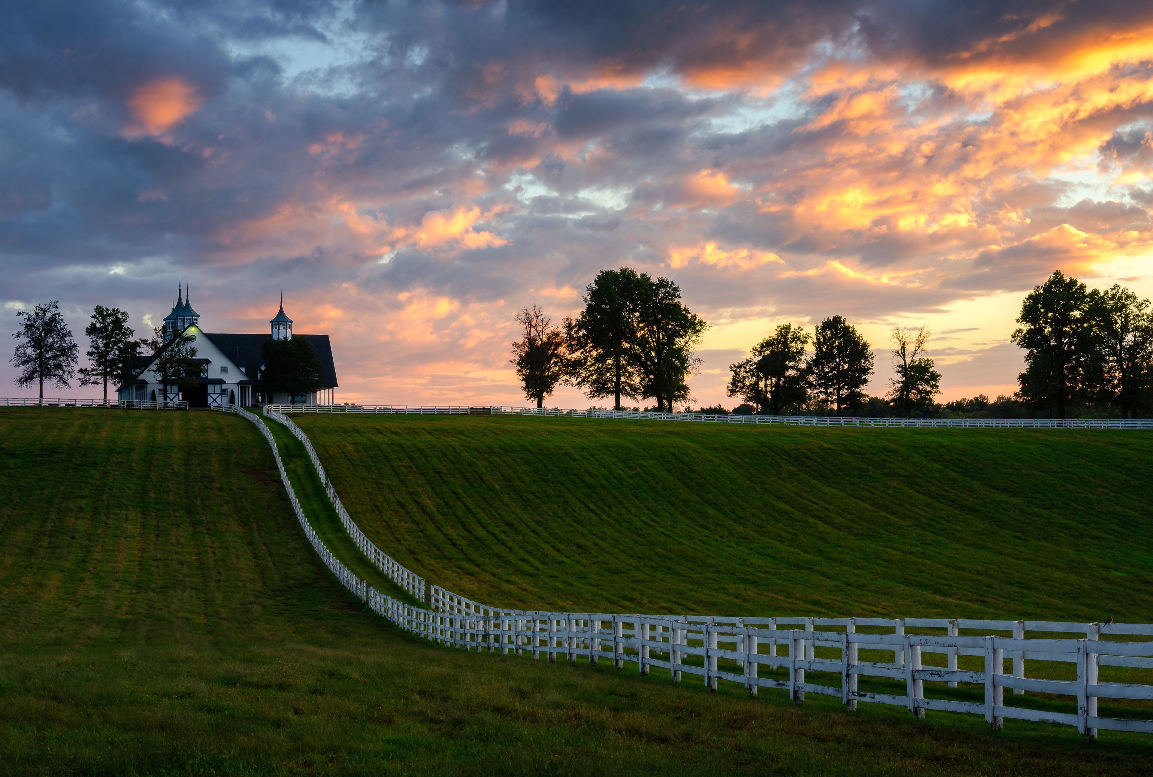 Paisaje rural con una cerca blanca, una iglesia y árboles bajo un cielo con colores de atardecer.