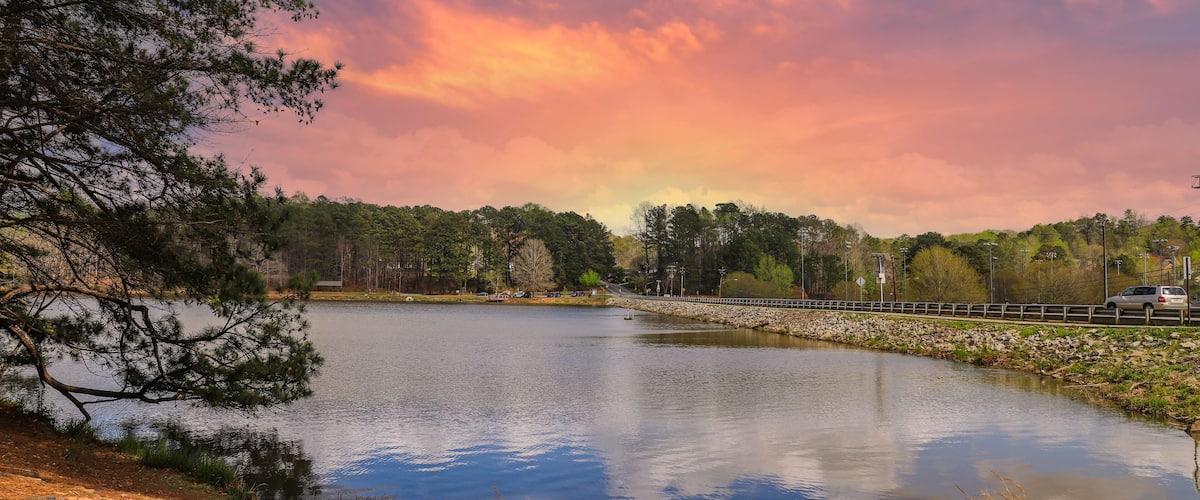 a stunning shot of the vast still waters of a lake surrounded by lush green trees with powerful clouds at sunset at Murphey Candler Park in Atlanta Georgia USA