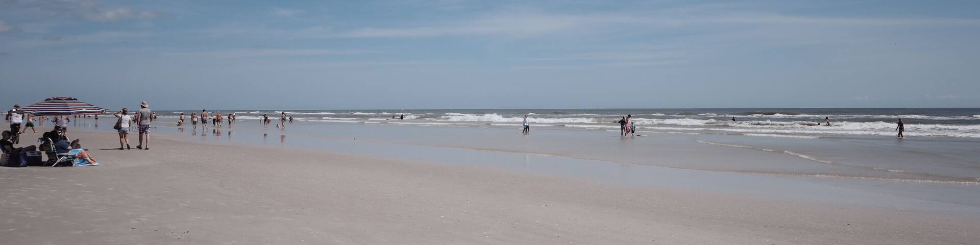 A beach with people walking and playing, a striped umbrella, and the ocean.