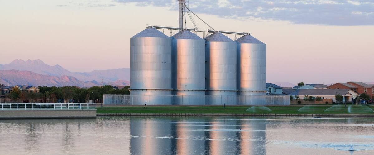 Four metal grain silos reflect early evening light beside a landscaped pond, illustrating the intersection of agricultural storage infrastructure and suburban development in a master-planned area