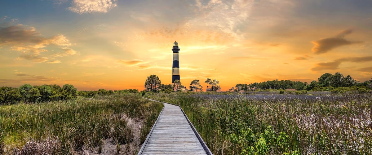 A lighthouse on a wooden boardwalk surrounded by tall grass and trees during sunset.
