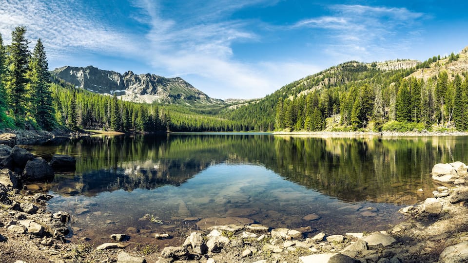 Panorama of a mountain lake with rocks in the foreground and trees in the far shore with mountains in the background, blue sky with high clouds. Strawberry Lake, Strawberry Wilderness, Oregon