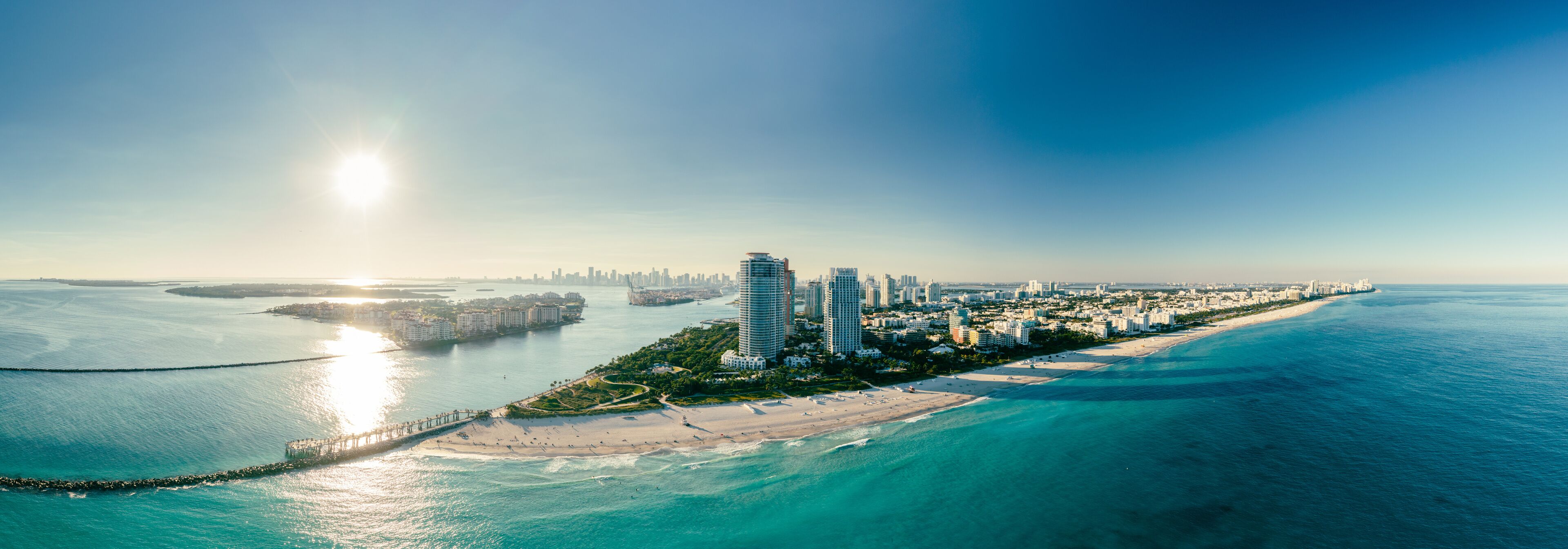 A coastal cityscape with a prominent pier extending into the sea, a sandy beach, and a dense cluster of high-rise buildings.