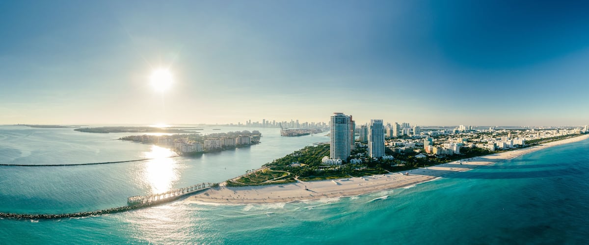 A coastal cityscape with a prominent pier extending into the sea, a sandy beach, and a dense cluster of high-rise buildings.