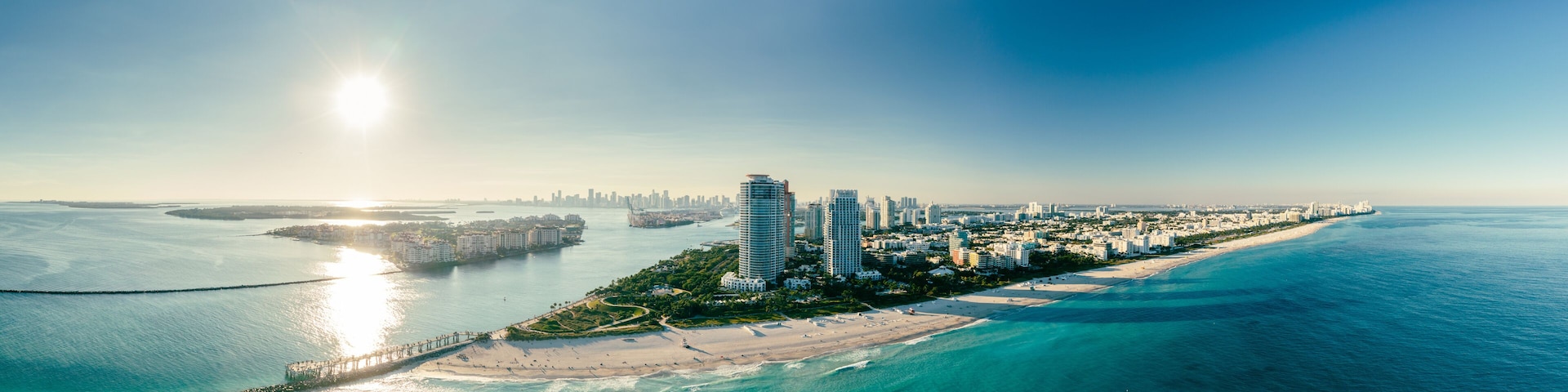 A coastal cityscape with a prominent pier extending into the sea, a sandy beach, and a dense cluster of high-rise buildings.