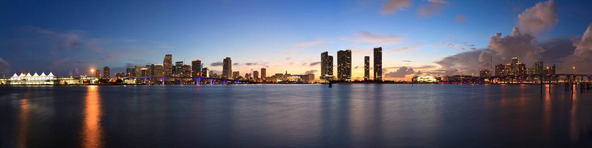 A city skyline at dusk with illuminated buildings and a calm river reflecting the lights.