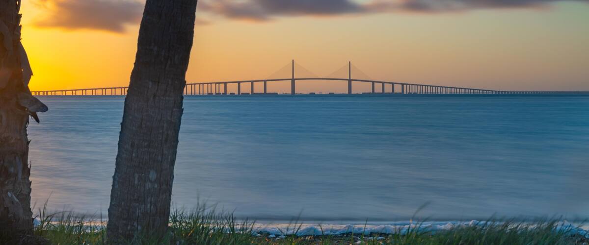This stunning photograph captures the Sunshine Skyway Bridge from Fort DeSoto Park at sunrise, where the first light of day casts a warm glow across the tranquil waters. The soft light the golden hr