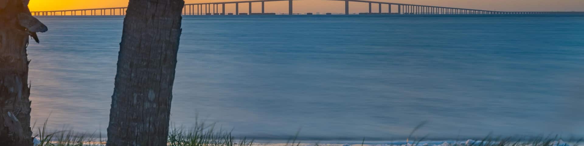 This stunning photograph captures the Sunshine Skyway Bridge from Fort DeSoto Park at sunrise, where the first light of day casts a warm glow across the tranquil waters. The soft light the golden hr