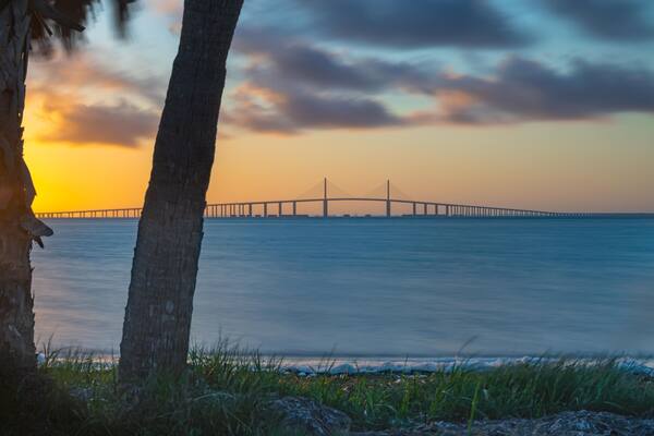This stunning photograph captures the Sunshine Skyway Bridge from Fort DeSoto Park at sunrise, where the first light of day casts a warm glow across the tranquil waters. The soft light the golden hr