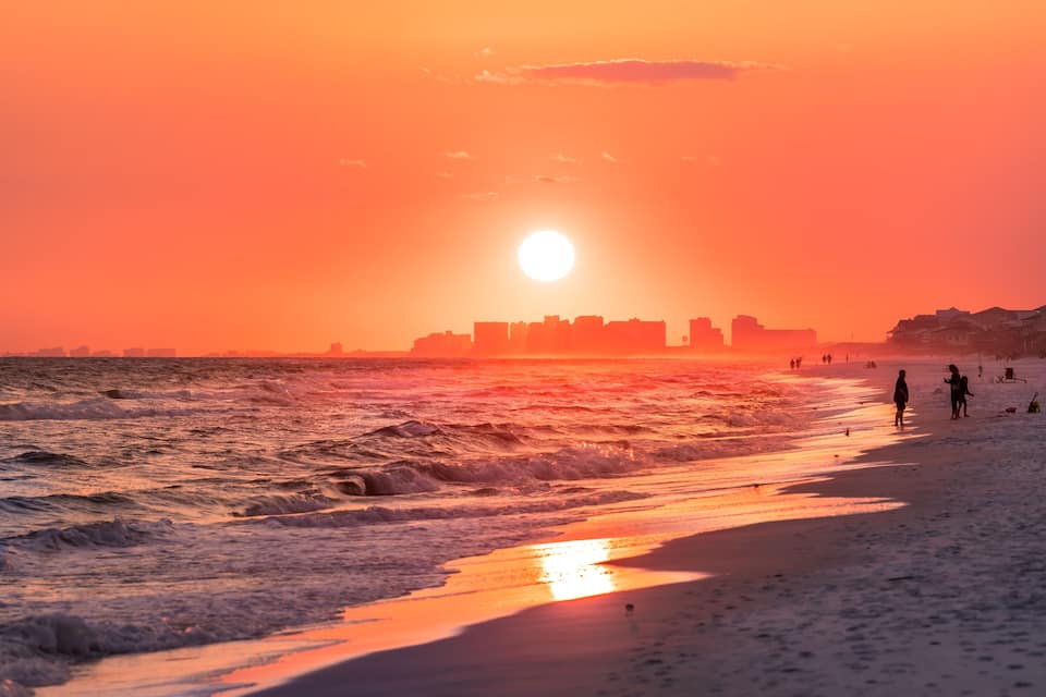 Ein Strand bei Sonnenuntergang mit Wellen, einer Stadtansicht in der Ferne und einigen Personen im Sand.