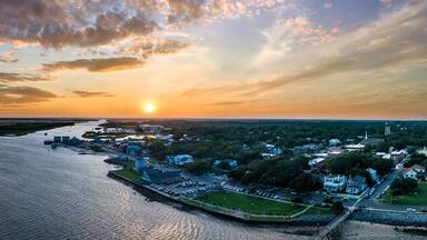 A coastal town at sunset with a river, waterfront buildings, and a lighthouse.