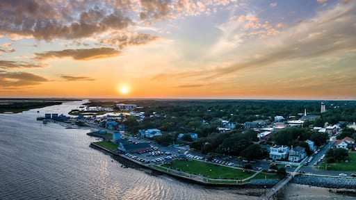 A coastal town at sunset with a river, waterfront buildings, and a lighthouse.
