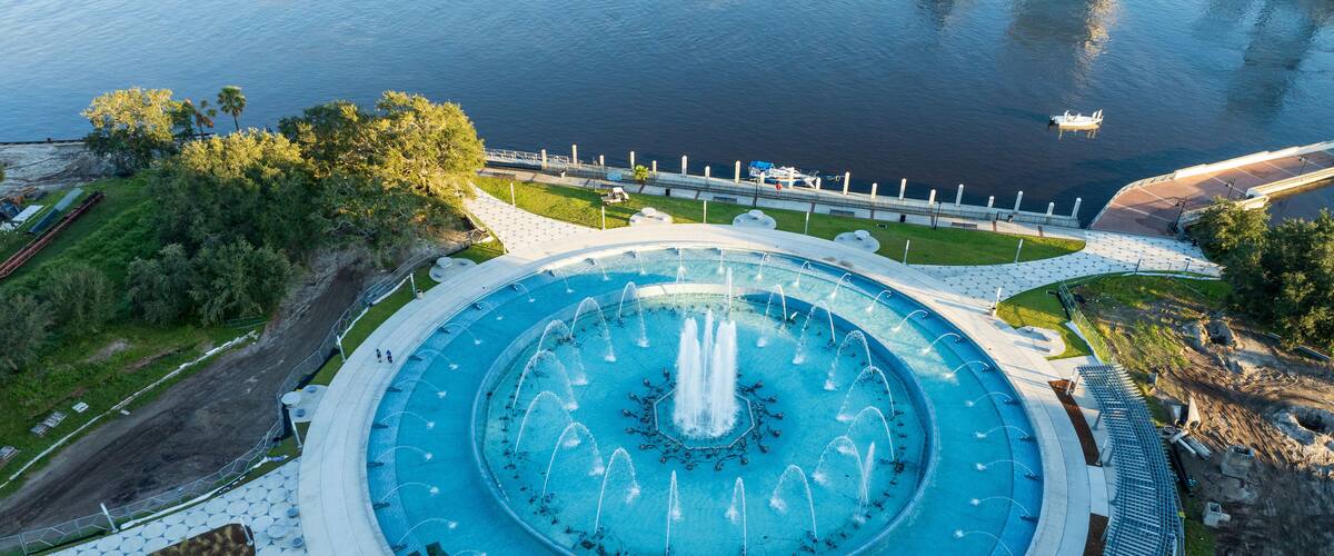 A large circular fountain with water jets in the center, surrounded by a paved walkway and greenery.