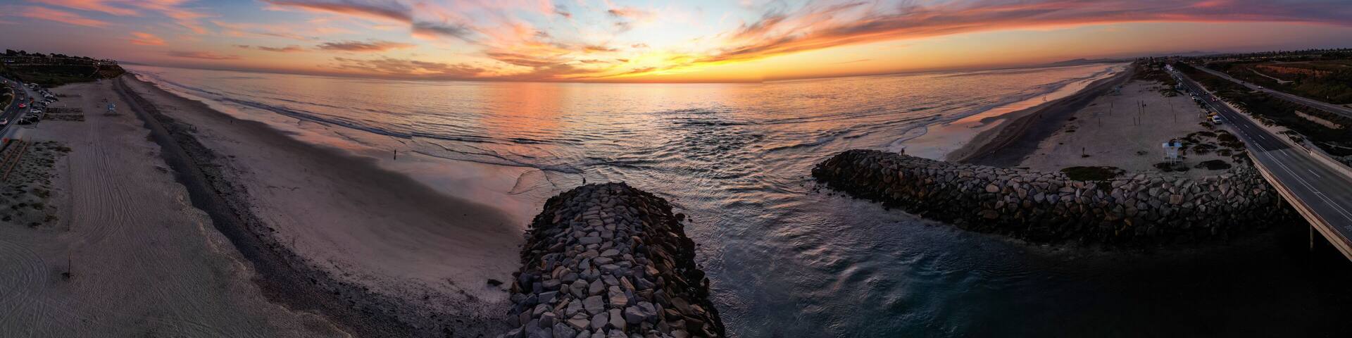 A coastal sunset with a stone jetty leading into the ocean, a road parallel to the shore, and a distant shoreline with buildings.