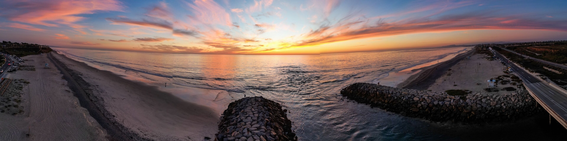 Um pôr do sol costeiro com um cais de pedra que se estende para o oceano, uma estrada paralela à costa e uma linha de costa distante com edifícios.