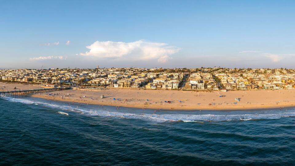 An aerial view of Manhattan Beach, California, highlights a sandy beach, a prominent pier, and a dense urban landscape under a clear blue sky.