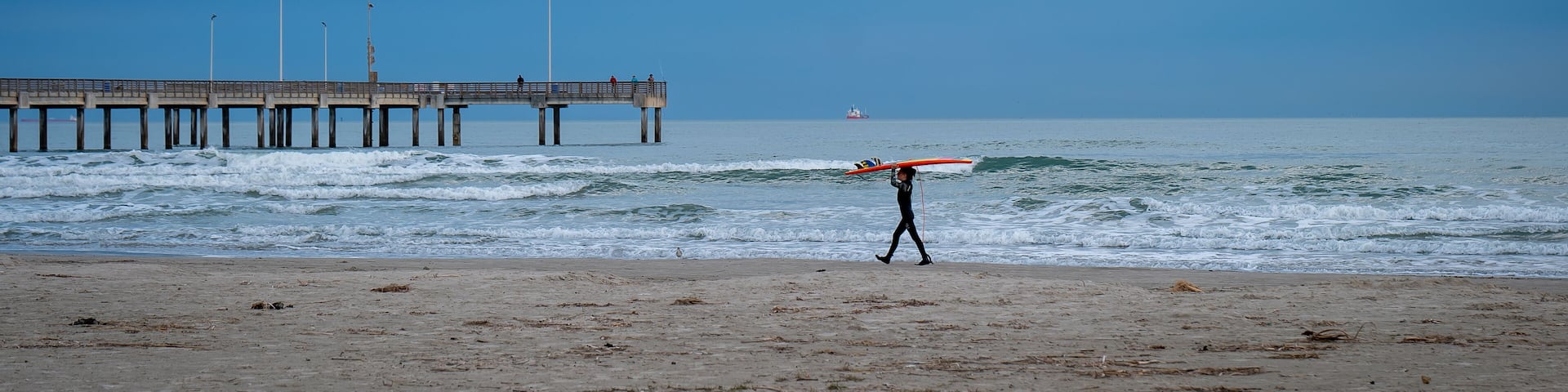 PORT ARANSAS, TX - 9 FEB 2023: Young surfer carries a surfboard on the beach, near the water of the Gulf of Mexico and a pier, in evening light.