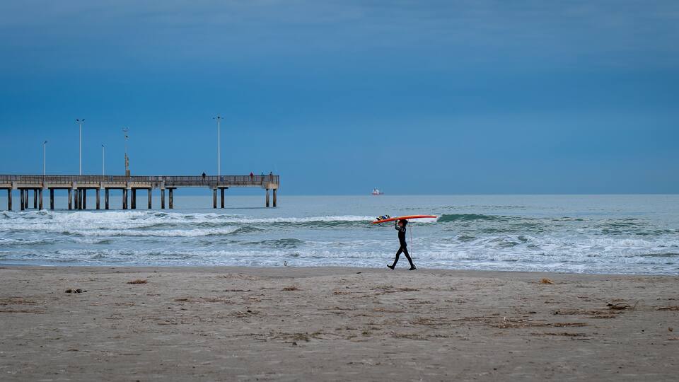 PORT ARANSAS, TX - 9 FEB 2023: Young surfer carries a surfboard on the beach, near the water of the Gulf of Mexico and a pier, in evening light.