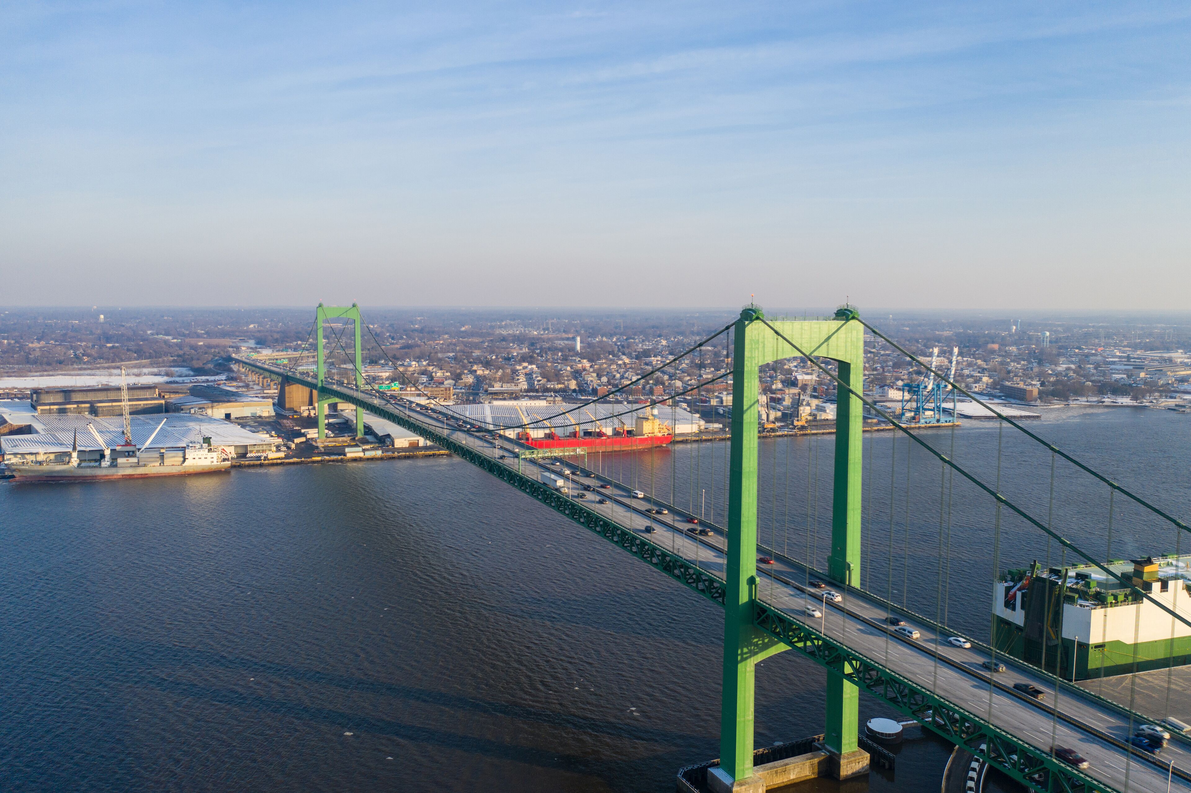 A green suspension bridge over a river with a cityscape in the background.