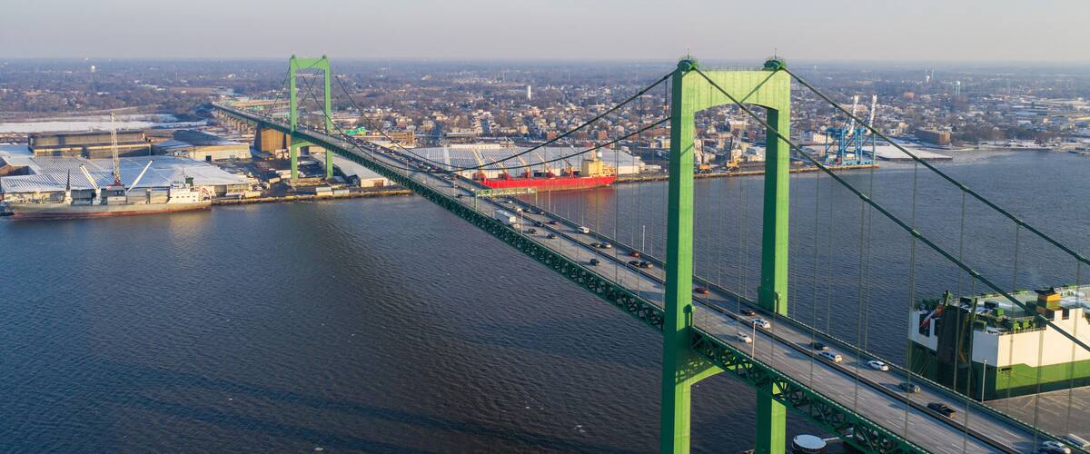 A green suspension bridge over a river with a cityscape in the background.