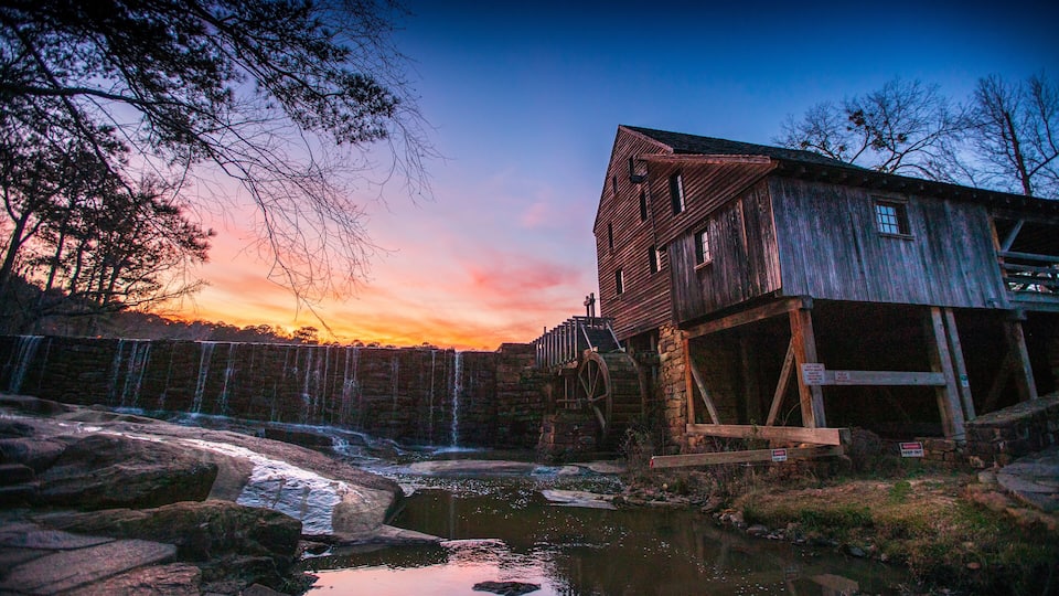 A rustic wooden mill with a water wheel, a small pond, and a sunset in the background.