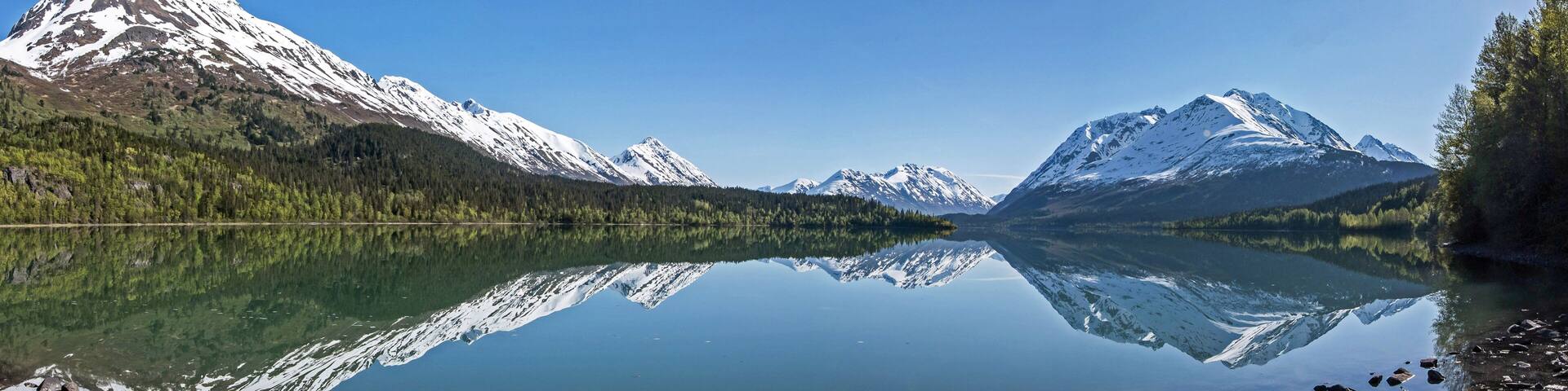 A calm lake reflecting snow-capped mountains, clear skies, and dense forest.