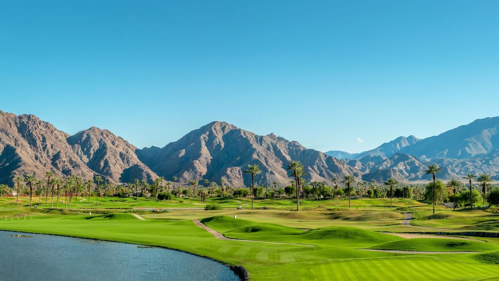 Un parcours de golf avec un lac, des palmiers et des montagnes en arrière-plan.