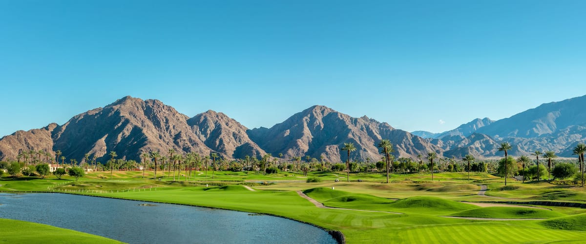 A golf course with a lake, palm trees, and mountains in the background.