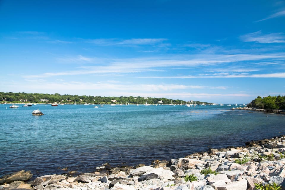 A coastal landscape with a rocky shoreline, calm sea, and several boats anchored in the distance.
