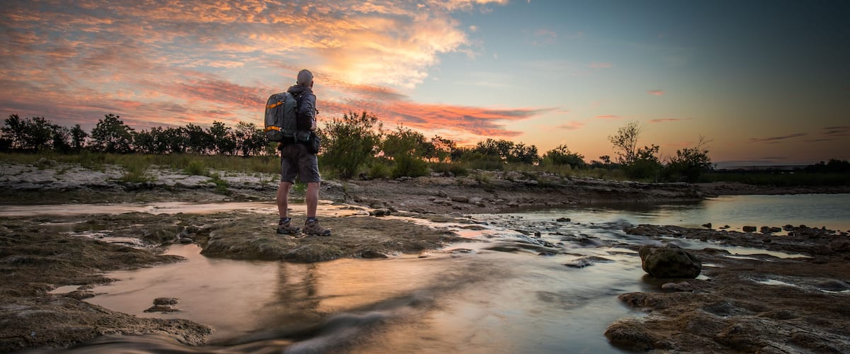 A person standing by a river at sunset with a backpack.