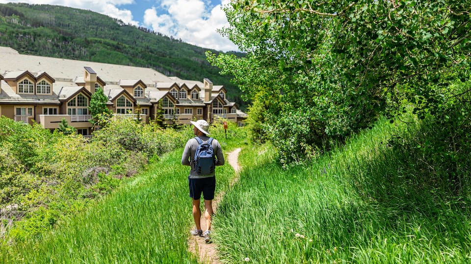 One young man back with backpack and hat walking on hiking trail of village loop in Beaver Creek ski resort near condo buildings in Colorado in summer
