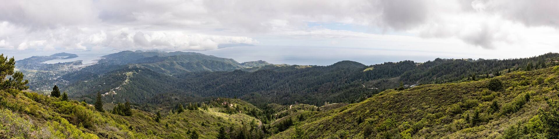 A mountainous landscape with dense forests, a winding road, and a distant body of water under a cloudy sky.