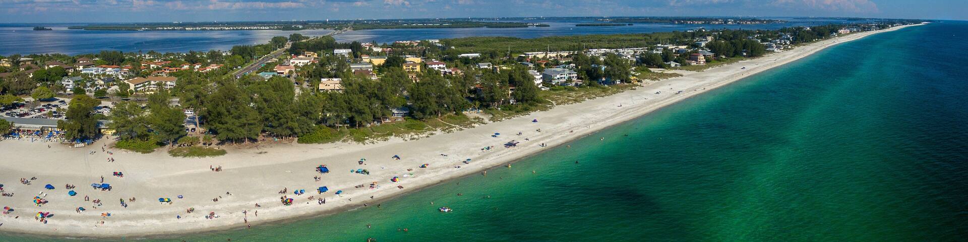 A coastal town with a long sandy beach, clear turquoise waters, and a backdrop of clouds.