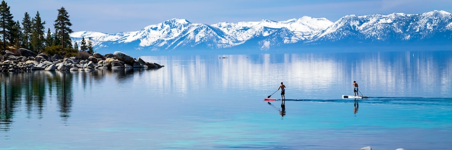 A calm lake with two paddleboarders, surrounded by snow-capped mountains and dense forest.