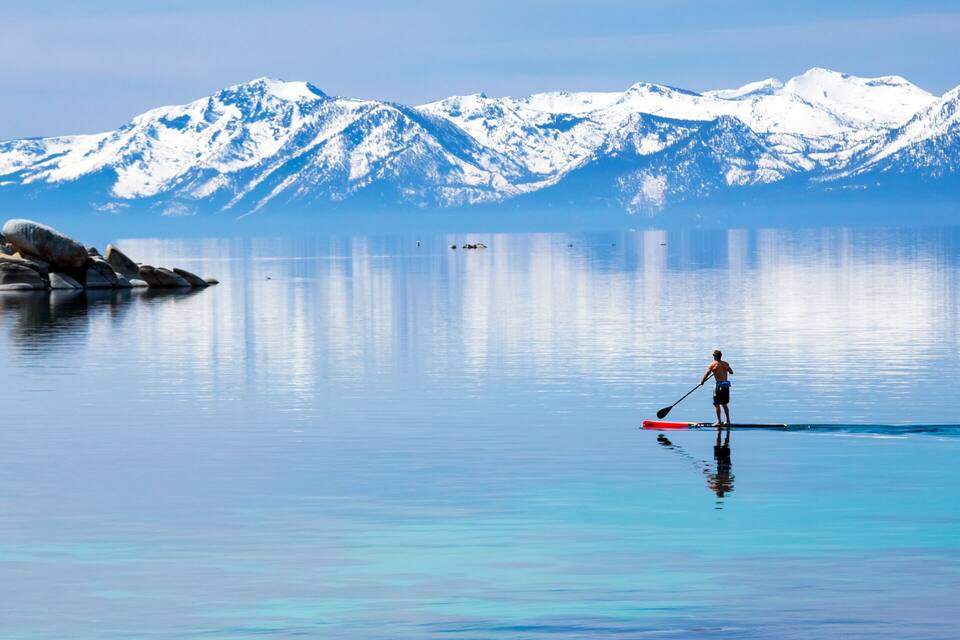 A calm lake with two paddleboarders, surrounded by snow-capped mountains and dense forest.