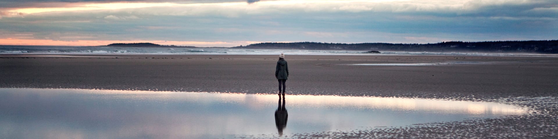 A person standing on a reflective wet beach with a cloudy sky.