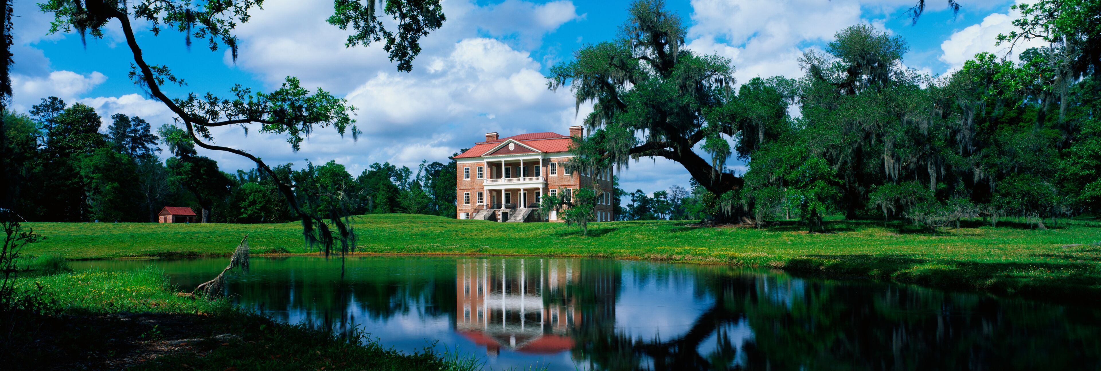 This is a southern plantation called Drayton Hall. It is a pre-Revolutionary plantation set on the Ashley River. It has Georgian Palladian architecture and was built from 1738-1742. It is set back on a green lawn with a pond showing the reflection of the plantation in the water.