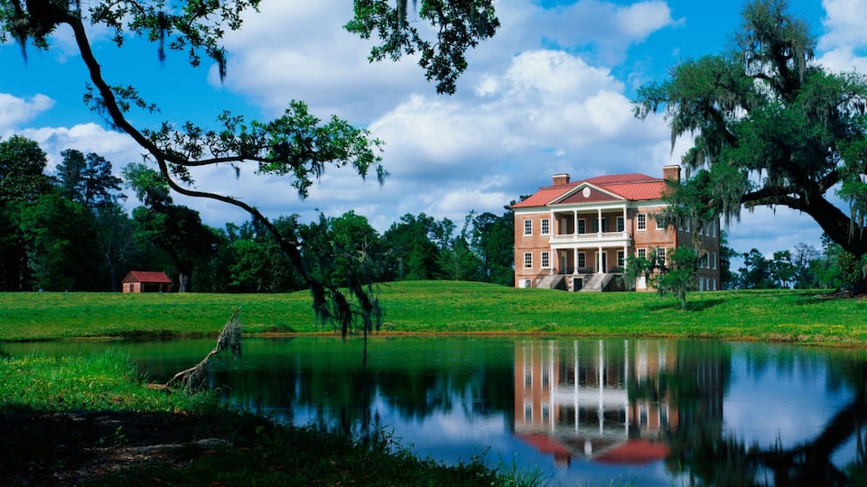 This is a southern plantation called Drayton Hall. It is a pre-Revolutionary plantation set on the Ashley River. It has Georgian Palladian architecture and was built from 1738-1742. It is set back on a green lawn with a pond showing the reflection of the plantation in the water.