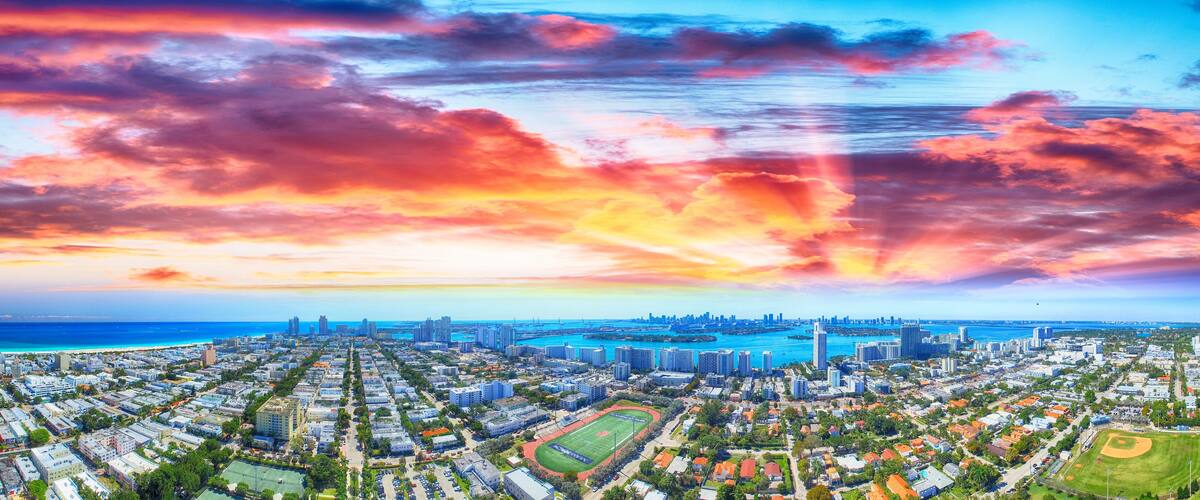A cityscape with a sunset over the ocean, featuring high-rise buildings, a sports field, and a marina.