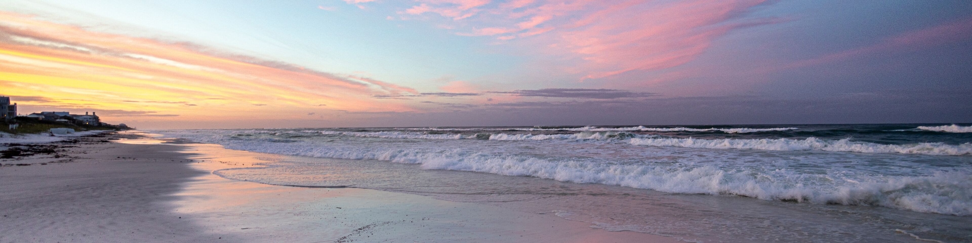 Uma praia com ondas ao pôr do sol, céu limpo e terra ao longe.