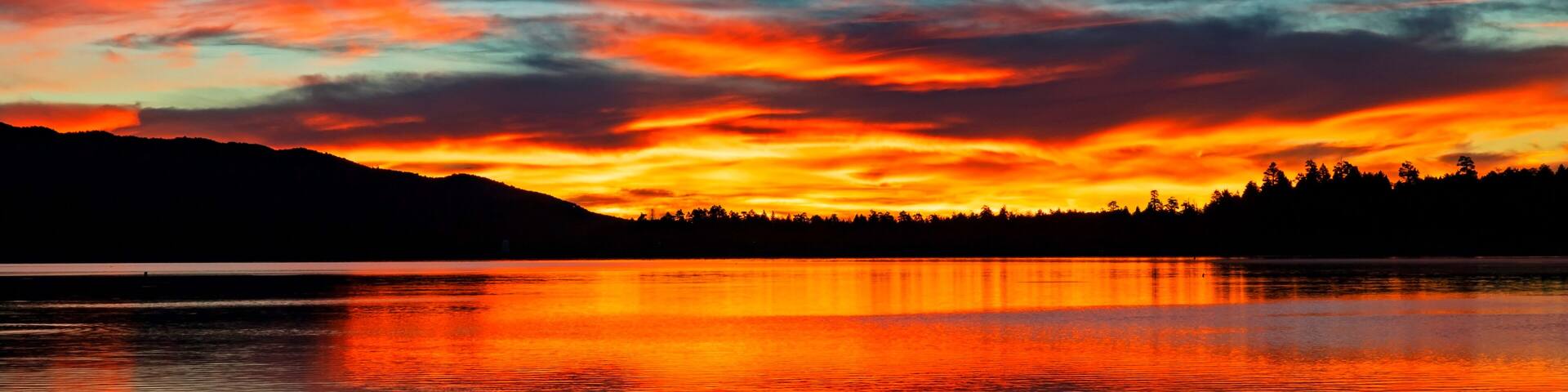 A sunset over a lake with vibrant red and orange clouds, calm water reflecting the colors, and a silhouette of a forested hill in the distance.