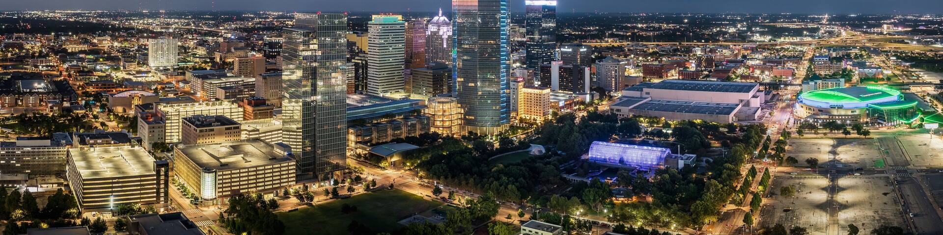 A cityscape at night with a prominent skyscraper, a stadium, and surrounding buildings.