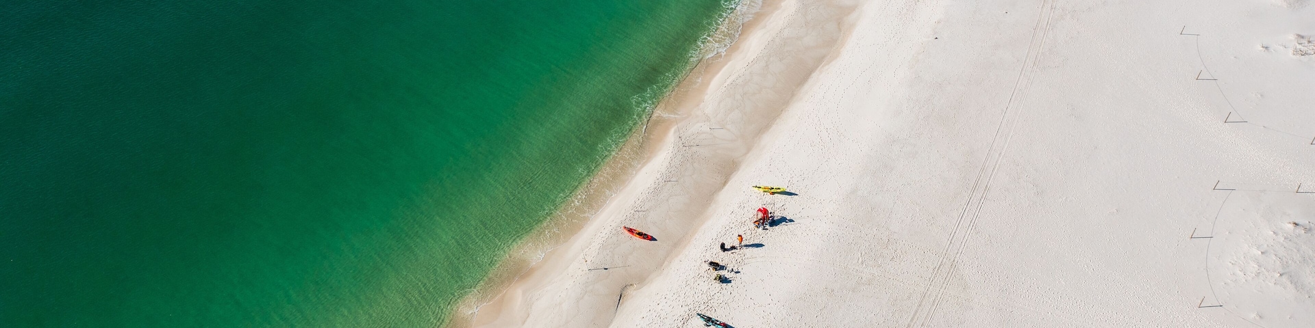 An aerial view of a beach with clear turquoise water, white sand, and a few people with colorful umbrellas.