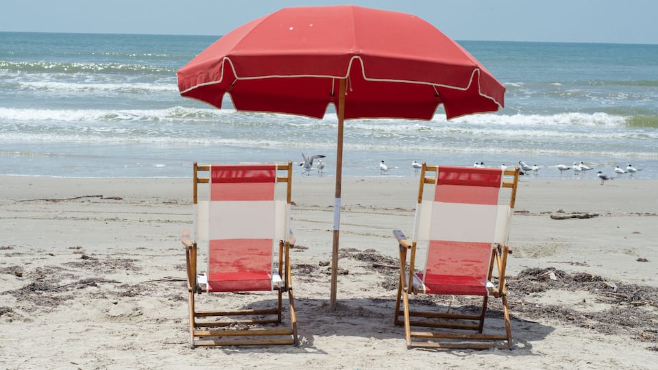 Two beach chairs with red cushions and a red beach umbrella on a sandy beach with the ocean in the background.