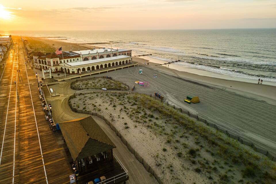 A beach with a boardwalk, a building with an American flag, and a sunset over the ocean.