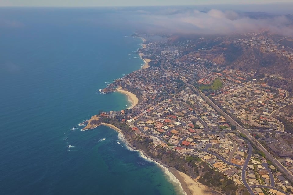 Vista aérea de uma cidade costeira com praia, área urbana densa e estradas sinuosas.